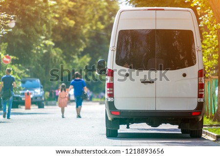 Back view of white passenger medium size commercial luxury minibus van parked n shadow of green tree on summer city street i with blurred silhouettes of pedestrians and cars under green trees.