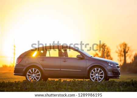 Modified image of a fictional non existent car. Gray car parked in countryside on blurred rural landscape and orange sky at sunset copy space background. Transportation, traveling concept.