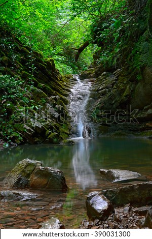 Beautiful landscape of waterfall in deep forest with big brown stones at foreground and tree branches background. Amazing view at fast water stream in tropical forest scenery and small green pond.
