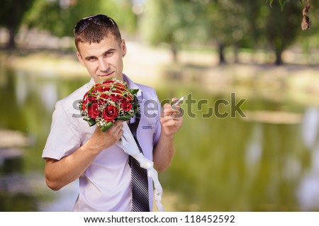 casual groom sniffing wedding bouquet of flowers. wedding. Lake shore. fiance smokes cigarette against the lake in a park. Love story of happy couple. Bridal wedding bouquet of flowers.
