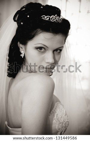 Young beautiful bride waits for groom near the window. Beautiful Bride lit by sunlight from a window. beautiful young bride standing beside a large window waiting and plays her veil