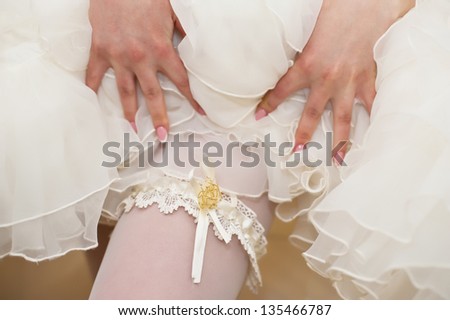 Garter on the leg of a bride, slim sexy bride in wedding luxury dress showing her silk garter with golden ribbon. woman have a final preparation for wedding ceremony. Wedding day moments