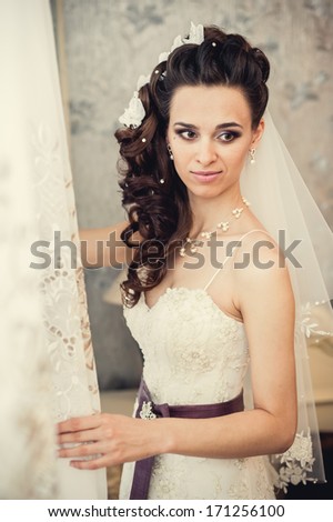 Young beautiful bride waits for groom near the window. Beautiful Bride lit by sunlight from a window. beautiful young bride standing beside a large window waiting