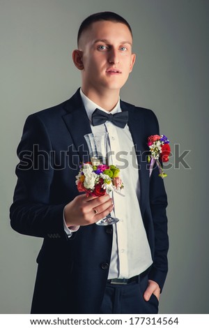 elegant young fashion man in tuxedo raising a glass of champagne. Portrait of stylish handsome man in classical suite and black bow tie. Standing and looking at camera isolated over gray background