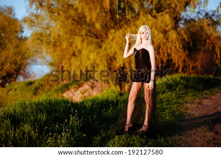 girl walking outdoor in black dress on the river bank at sunset. Beautiful blonde model in long black dress standing on the green grass near the beach.