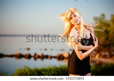 girl walking outdoor in black dress on the river bank at sunset. Beautiful blonde model in long black dress standing on the green grass near the beach.