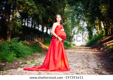 Pregnant woman in red dress outdoor. The young pregnant woman on walk in park. Beautiful healthy pregnant woman