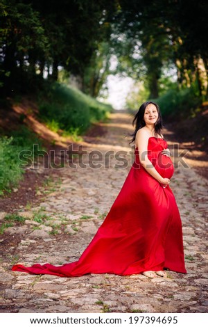 Pregnant woman in red dress outdoor. The young pregnant woman on walk in park. Beautiful healthy pregnant woman