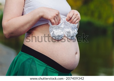 Belly of a pregnant woman on the nature. Girl holds small baby slippers. Beautiful belly of young attractive pregnant woman walking outdoor