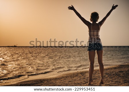Beautiful young woman on the beach holding hands above the head. warm summer evening sunshine. arid landscape. Happy Woman in a short dress enjoys in Sea Sunset. In the background sea, vintage color