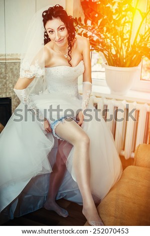 Garter on the leg of a bride, slim sexy bride in wedding luxury dress showing her silk garter with golden ribbon. woman have a final preparation for wedding ceremony. Wedding day moments