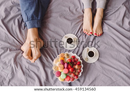 Young caucasian couple having romantic breakfast in bed. Closeup of female and male feet, two cups of coffee, fruits and colorful biscuits.