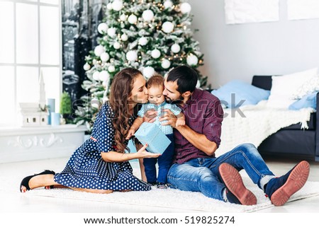 Modern Christmas Family Portrait In Home Holiday Living Room. Mother and father kissing Kid Baby waiting Santa With Present Gift Box, House Decorating By white Xmas Tree Candles Garland