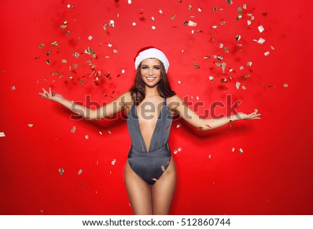 beautiful young girl model stands on red background in sexy bathing suit  bikini and a Christmas Santa hat.on the background of flying golden candy.advertising photo. Summer vs. winter.New Year at Sea
