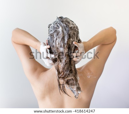 The back of a woman washing and shampooing her hair full of suds in front of a white background.