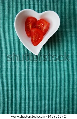 Red tomatoes salad in heart shape bowl on green background, free space for text