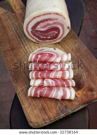 Pancetta, typical italian roll raw bacon with slices on wood trencher