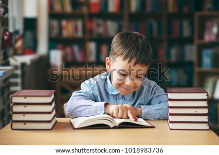 Preschool european boy is staying at the table in the library with stacks of books beside. Close-up pupil holding his hands on a book while reading. Pupil loves lecture, preparing for school.