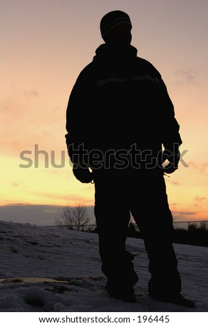 Silhouette of man in snow gear standing on hill beside snow board.