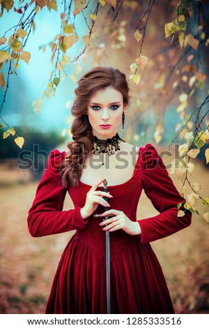 Portrait of magnificent Fashion gothic girl standing in autumn forest .Fantasy art work.Amazing red haired model in claret dress with a sword .Fairytale about young princess-warrior.