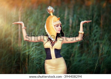 Art work Egyptian Queen Cleopatra. Shooting outdoors in the style of ancient Egypt. A beautiful young girl in gold dress and Egyptian head wear raising hands in sun light. Egyptian make up face.