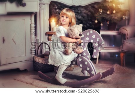 Adorable little blonde girl sitting on a toy horse near christmas tree and holding a toy bear. Pretty kid in a warm cozy room. New year photo. Art work.