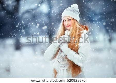 Beautiful girl with long curly hair in  white knitted scarf and hat having fun outdoor in winter forest under snowflakes. Pretty young model standing  on blue background and looking at camera.