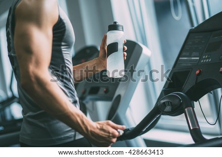 Unrecognizable young man in sportswear running on treadmill at gym and holding bottle of water