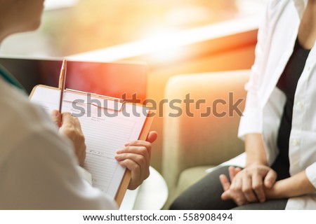Professional medical doctor in white uniform gown coat interview counseling female patient: Physician writing on patient chart while consultation: Hospital/ clinic healthcare professionalism concept