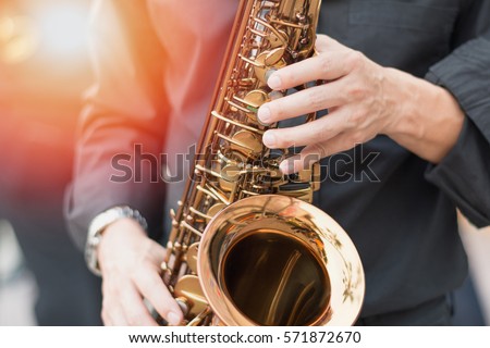 Closeup hands of man playing golden alto saxophone with light effect. Image of musician male player in black shirt blowing music instrument in outdoor park jazz music festival.