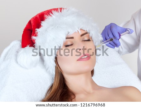 The cosmetologist makes the Botulinum toxin injection on the face skin of a beautiful, young woman in the Santa Claus hat. New Year\'s and Cosmetology concept.