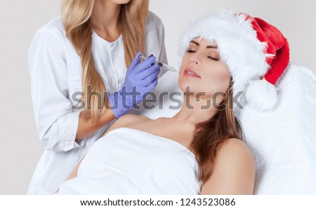 The doctor cosmetologist makes  injection on the face skin and lips of a beautiful, young woman in the Santa Claus hat. New Year\'s and Cosmetology concept.