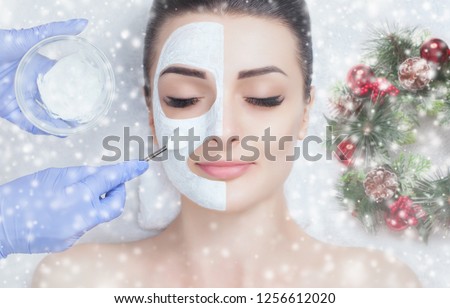 The cosmetologist for the procedure of cleansing and moisturizing the skin, applying a mask with stick to the face of a woman. Closeup woman face and new year wreath with snowflakes.