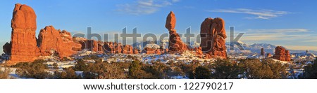 Panorama of landmark Balanced Rock and other snowy red rock pillars against the backdrop of the snow-capped La Sal Mountains in Arches National Park, Utah