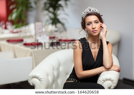 Beautiful girl  in a black suit with a tiara on her head sits in a chair