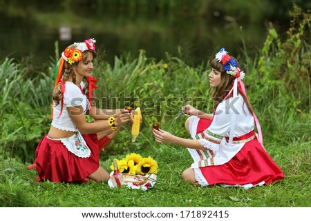 Two girls in Ukrainian costumes dressed for the holiday of Ivan Kupala sitting on the Bank of the river and drink vodka