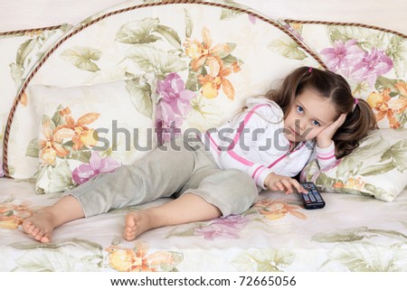 The little girl lies on a sofa and switches channels of the TV by means of a remote control