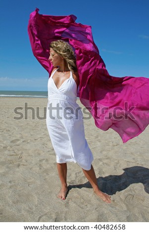 young woman in white dress on ocean beach with red scarf fluttering on wind