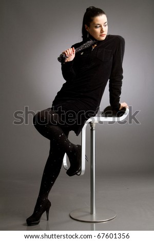 Beautiful woman with long blach hair and in black chlotes sitting on bar stool on gray background