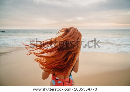 young red-haired woman with flying hair on the ocean coast at sunset, rear view