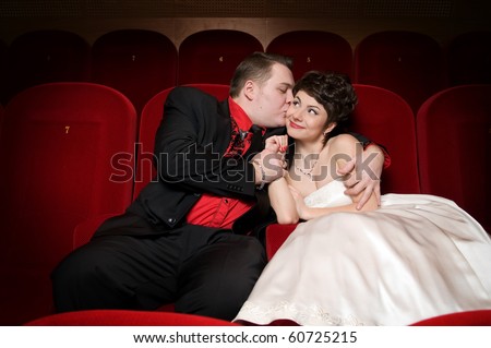 In a cinema hall the guy in a red shirt embraces and kisses the beautiful girl in a red dress on a cheek