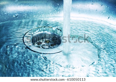 A stream of clean water flows into the stainless steel sink in blue tones. Sink plug hole close up macro .