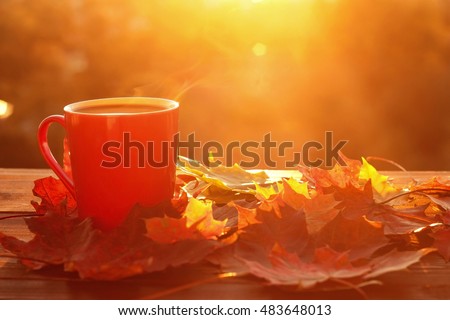 Autumn leaves and hot steaming cup of coffee. Wooden table against golden sunset or sunrise light background. Fall season, leisure time and coffee break concept.
