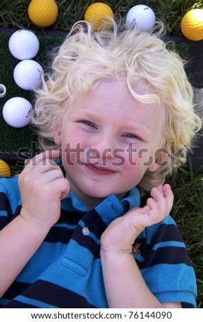 Beautiful smiling blond boy laying on grass surrounded by white and yellow golf balls