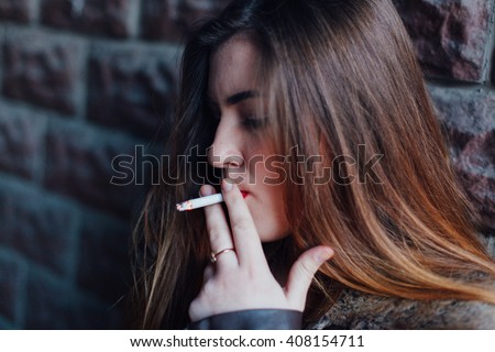 Portrait of a beautiful woman smoking in a warm leather jacket leaning on the loft brick wall, smokes under stress, wanting to relax, letting out smoke cigarettes