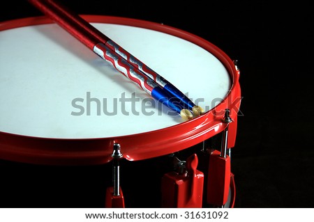 A red and black colored snare drum with flag colored sticks against a black background in the horizontal format.