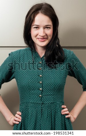 Young female model with a natural look smiling while having her hands on hips, She\'s wearing a green dress with white dots.