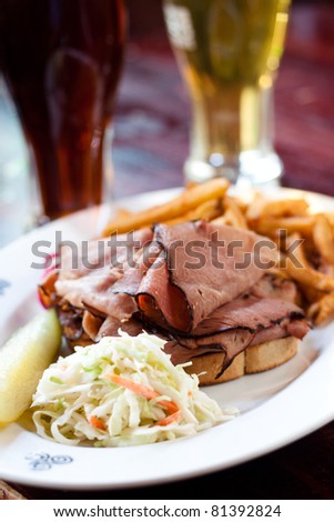One half pound of steamed and shaved Montreal smoked meat piled high on fresh rye bread. Served with creamy coleslaw, fries and pickle. Pints of beer in the background.