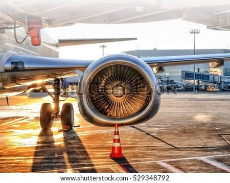 Modern passenger airplane parked to terminal building gate at airside apron of airport with airplane parts jet engine wing landing light windows gear sunset setting sun view air travel background