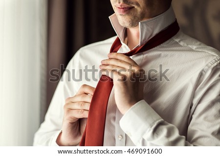 People, business, fashion and clothing concept - close up of man in shirt dressing up and adjusting tie on neck at home. Businessman putting on a tie. Man putting on necktie. Dressed in the morning